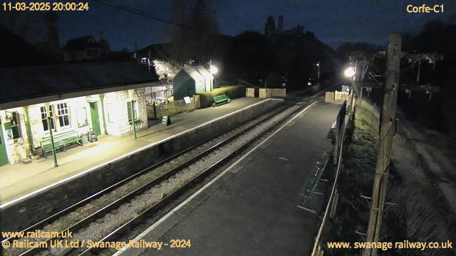 A dimly lit railway station at night, featuring a stone platform with two sets of railway tracks. On the left, there is a building with large windows and a green bench in front. A green sign that says "WAY OUT" is visible. The right side of the image shows a wooden fence with additional green benches and a lamp illuminating the area, while a tall wooden utility pole stands next to the tracks. There is sparse vegetation and no visible people.