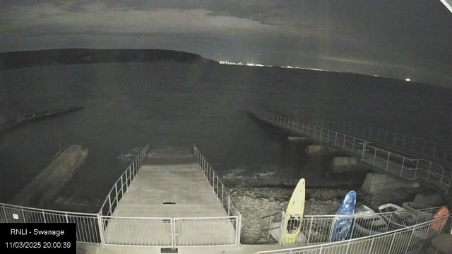 The image shows a dark waterfront scene at night. In the foreground, there is a concrete ramp leading down to the water, bordered by a metal railing. To the right, two kayaks are positioned on the ground, one yellow and one blue, next to some scattered equipment. In the background, the water appears calm but dark, with a distant coastline visible against the night sky, faintly lit by lights. The sky is mostly cloudy, contributing to the dim lighting of the scene.