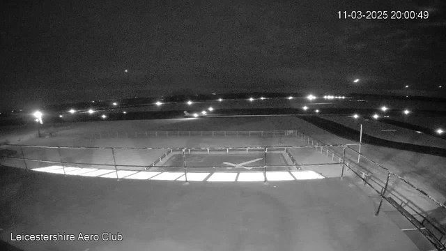 A nighttime view from a webcam at Leicestershire Aero Club. The foreground features a flat, dark rooftop with a railing. In the center, a lit area with a faint cross shape is visible. In the background, the horizon is dotted with lights from distant buildings or vehicles. The sky is mostly dark with a few scattered clouds. The time displayed on the image is 20:00:49 on March 11, 2025.