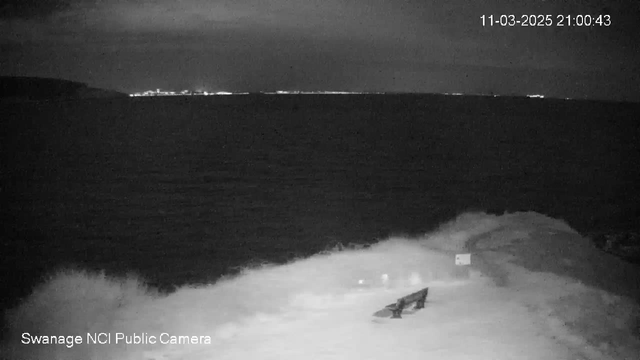 A night scene captured by a webcam shows the ocean with waves crashing against a rocky shoreline. In the foreground, there is a small wooden bench. The background displays a distant city skyline illuminated by lights, contrasting with the dark water. The sky appears overcast with clouds, making the scene predominantly dark and moody. A timestamp in the corner indicates the date and time of the image.