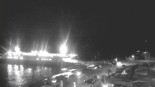 A nighttime scene featuring a brightly lit ferry docked at a pier, casting reflections on the water. The foreground shows a road lined with parked cars and a few illuminated areas, including streetlights and nearby buildings. The sky is dark, with minimal visibility beyond the lights.