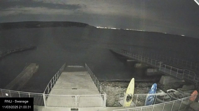 A dark shoreline scene with a calm sea under a cloudy sky. A wooden dock extends into the water, with a smaller jetty visible on the right. Two kayaks, one yellow and one blue, are resting on stony ground near the dock. The distant outline of a hill can be seen on the horizon, with faint lights indicating a town or city. The overall atmosphere is quiet and night-like, with limited visibility.