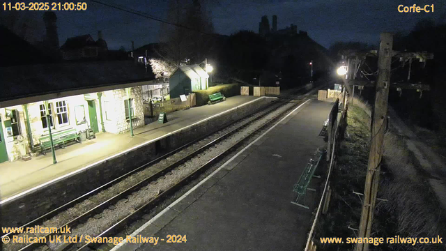 A dimly lit railway station is shown at night. The platform is empty, with several green benches lining the edge. A low stone wall separates the platform from the railway tracks, which are visible in the foreground. To the left, there is a building with stone walls and large windows, illuminated by warm light. A sign labeled "WAY OUT" can be seen nearby. A distant wooden fence is positioned at the end of the platform, along with a wooden pole bearing wires and a light. The overall scene captures a quiet, peaceful atmosphere.