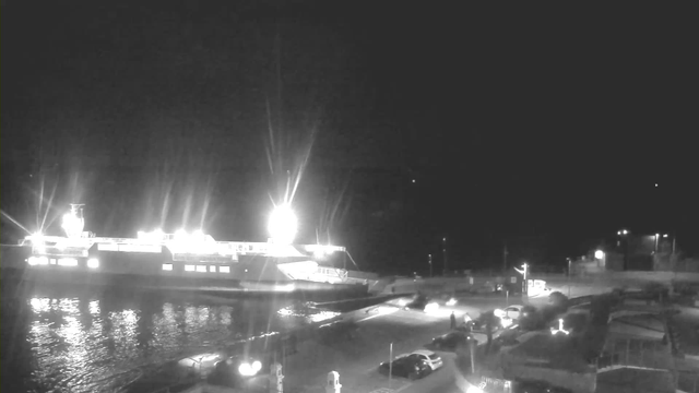 A dimly lit scene featuring a large ferry docked at a harbor, illuminated by bright lights from the vessel and surrounding area. The water reflects the lights in a shimmering pattern. In the foreground, there is a parking lot with several vehicles and scattered streetlights, while pedestrians can be seen walking along the shore. The sky appears dark, suggesting it is night time.