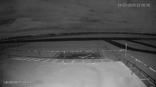 A dark, nighttime scene from a webcam at the Leicestershire Aero Club. The foreground features a flat surface with a railing and a section marked for helicopter landing, indicated by an "X" shape. In the background, faint outlines of distant lights are visible against a cloudy sky. The image has a monochrome, low-light quality, emphasizing the dark surroundings. The timestamp in the corner shows the date and time as 11-03-2025, 22:00:30.