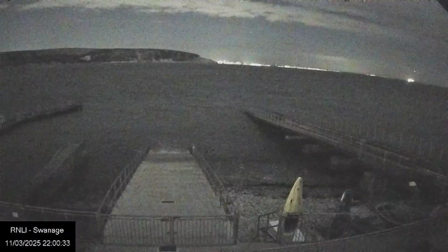 A dimly lit scene depicting a water body, likely the sea, under a cloudy night sky. In the foreground, there is a wide concrete platform leading to the water, with a railing surrounding it. To the right, there is a yellow object that resembles a kayak or small boat. A wooden jetty extends from the left side of the image into the water. In the background, faint lights from the opposite shore can be seen, creating a subtle horizon where the sea meets the sky.