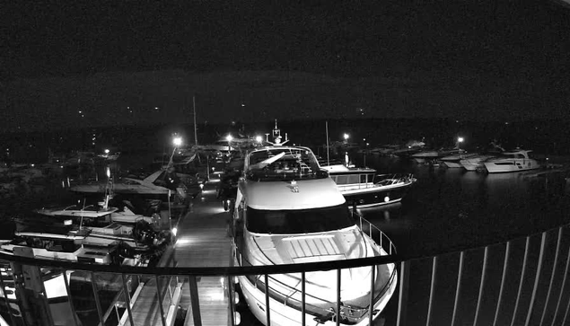 A dimly lit marina scene at night, featuring several boats and yachts lined up along a wooden dock. Some boats are illuminated by lights, creating reflections on the dark water. The overall atmosphere is calm and serene, with a few distant lights visible in the background.