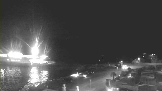 A nighttime scene captured from a webcam showing a harbor. On the left side, a brightly lit ferry is docked, illuminating the water with reflections. The surrounding area is dark, with some light coming from street lamps. In the foreground, there are a few parked cars and part of the harbor's edge, while the background includes a faint silhouette of buildings. The overall atmosphere is calm and quiet, with the harbor waters appearing still.