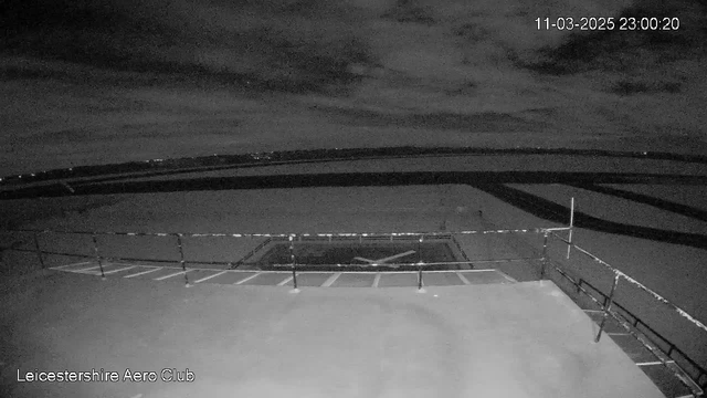 A night view from a webcam at Leicestershire Aero Club shows a flat, open area with a large outlined helipad in the center. The surface is somewhat illuminated, revealing a landing area and several lines marking the boundaries. In the background, faint lights are visible, possibly indicating buildings or vehicles far away. The sky is dark with scattered clouds, creating a mostly shadowy scene with little visible detail.