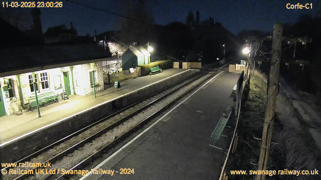 A dimly lit railway station at night. The scene shows a quiet platform with stone walls and a wooden bench. There are green benches against the wall and a sign indicating "Way Out." The platform is bordered by wooden fencing, and on the right side, there’s a telephone pole with wires. The railway tracks run along the foreground. In the background, some buildings are faintly visible, partially illuminated.