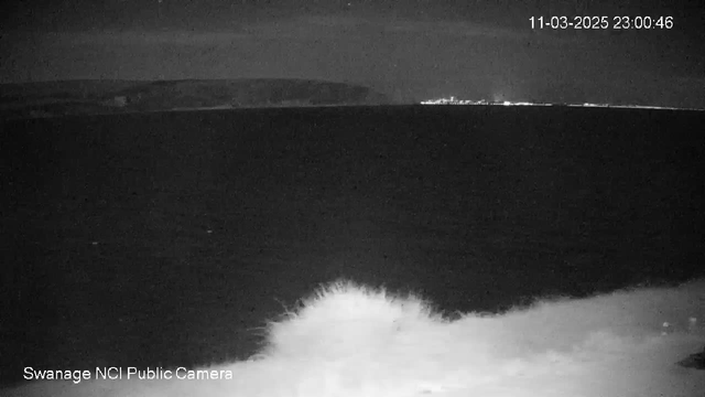 A black and white image showing a view of the sea under a night sky. The coastline is visible in the background, with a distant light possibly indicating a city or town. The foreground features a splash of white waves crashing against the shore. The image timestamps the date and time in the top right corner.