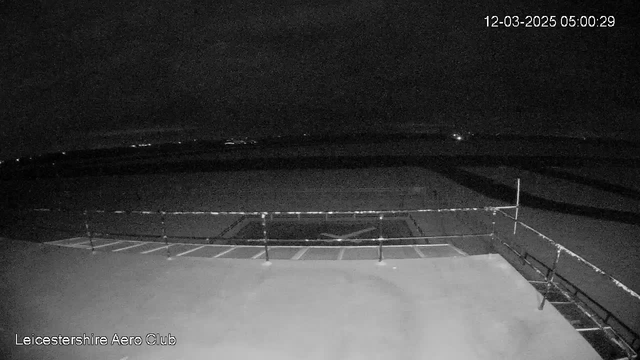 A dark, nighttime view from a webcam located at the Leicestershire Aero Club. The image shows an empty, flat surface with faint grid lines, indicating a runway or area for aircraft. In the background, there are distant lights twinkling on the horizon, suggesting the presence of buildings or vehicles far away. A railing is visible in the foreground along the left side of the image, and there are no clouds in the sky. The time stamp in the top right corner reads 05:00:29 on March 12, 2025.