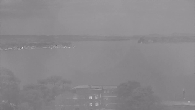 A foggy, monochrome view of a lake with distant land and buildings along the shoreline. In the foreground, there is a structure with several windows, while trees are partially visible. The background features a hazy horizon where the water meets the sky, and a small island can be seen in the distance.