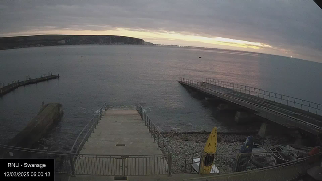 A view of a calm sea under a cloudy sky at dawn. The horizon shows a lightening sky with hues of orange and yellow. In the foreground, there is a concrete ramp leading down to the water, flanked by metal railings. To the right, a wooden jetty extends into the sea, and a yellow kayak is on the pebbled shore near some other boats partially visible. The landscape in the distance features cliffs and a coastal area, slightly illuminated by the early morning light.