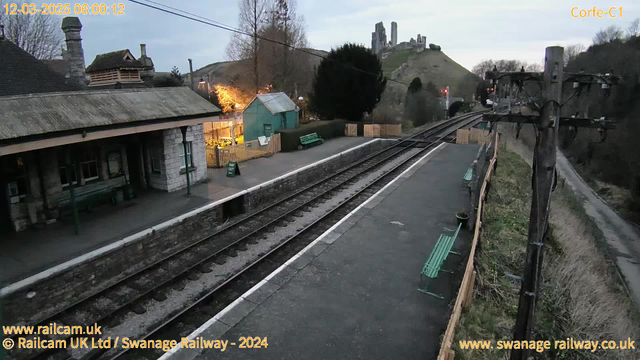 A train station scene during early morning light. In the foreground, there are two sets of train tracks running horizontally, with a section of the platform visible. To the left, a stone building with a sloped roof features large windows and wooden benches outside, surrounded by trees and lit with warm lights. On the right, a wooden fence separates the platform from a grassy area. In the background, a hill rises with the remains of a castle or ruins. Electric poles line the side of the tracks, with a visible signal light. The sky is overcast, and the overall mood is calm and quiet.