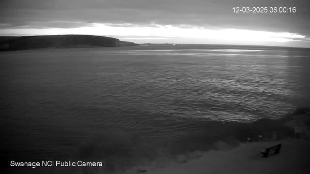 A black and white image of a calm sea under a cloudy sky at dawn. The horizon is slightly illuminated by the emerging light. A rocky shoreline is visible on the left side, and a wooden bench sits on the beach in the foreground. The scene is tranquil, with gentle ripples on the water’s surface. The time displayed at the top right is 06:00:16 on December 3, 2025.