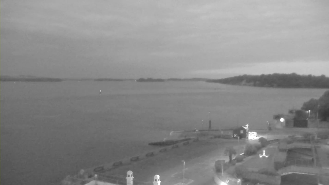 A black-and-white image showing a calm body of water under a cloudy sky. In the foreground, there are structures resembling lighthouse towers with rounded tops. A pier extends into the water with a few small boats moored along the side. In the distance, outlines of land can be seen on the horizon, partially obscured by the clouds. The overall mood is serene and atmospheric, with low lighting illuminating certain areas.