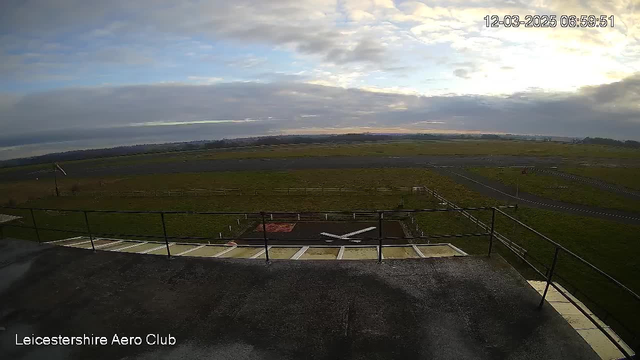 An outdoor scene captured from a webcam at Leicestershire Aero Club, showing a wide expanse of grassy airfield under a cloudy sky. In the foreground, there is a railing, and a grassy area with some markings and a red patch on the ground. The airfield extends into the distance where it meets a line of low hills against the cloudy sky. The timestamp at the top right indicates the date and time of the capture.