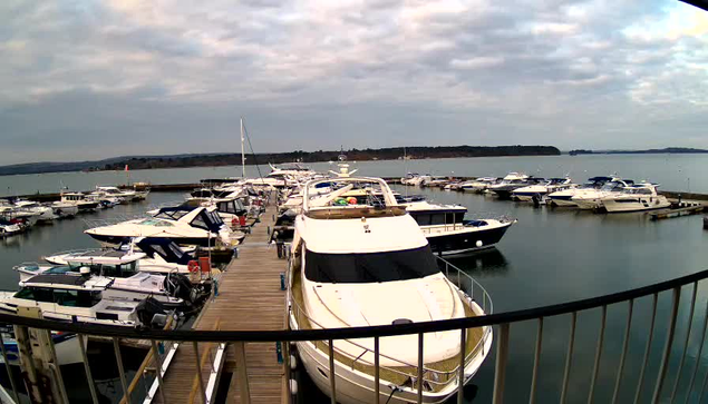 A view of a marina filled with various boats and yachts, some docked along wooden piers. The water is calm, reflecting the cloudy sky above. In the distance, there are hills or land along the horizon. The scene conveys a tranquil waterfront atmosphere.