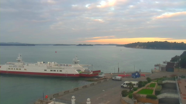 A calm waterfront scene at sunset featuring a ferry docked at a concrete pier. The ferry is white with red accents, displaying signs about speed and navigation. In the background, the water is serene, reflecting the light of the setting sun, and distant islands are visible on the horizon. The sky is partly cloudy, with hues of orange and pink near the sun. There are some vehicles and structures on the shore, including a small blue building and greenery in the foreground.