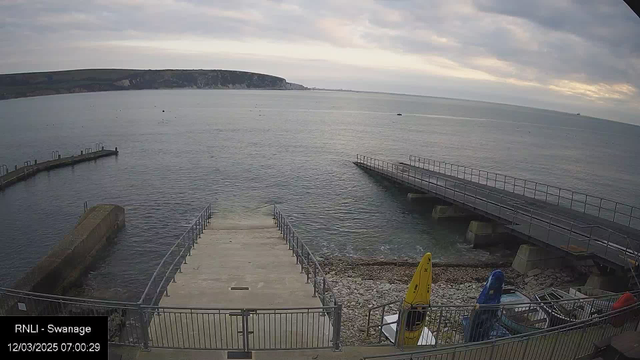 A coastal scene featuring a calm, reflective sea under a cloudy sky. On the left, there is a concrete ramp leading down into the water, with a metal railing along the sides. To the right, a wooden jetty extends over the water, with another ramp that leads down. In the foreground, there are several brightly colored boats, including yellow and blue, secured on the shore. The background shows a rocky shoreline and a distant cliff, partially obscured by clouds.