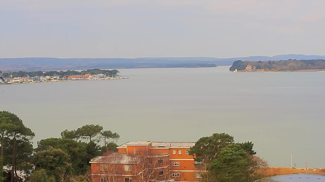 A panoramic view of a calm water body, with a shoreline lined with small buildings, boats, and trees. In the foreground, there is a red brick building with several windows, partially obscured by green foliage. The water reflects a muted sky, with rolling hills visible in the background, creating a serene landscape.