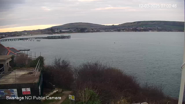 A coastal view at sunrise with calm water in the foreground. To the left is a wooden pier extending into the sea. The shoreline features buildings in the distance, surrounded by rolling hills that rise gently behind them. The sky is mostly cloudy with patches of light, indicating early morning. In the foreground, there are shrubs and a partially visible structure with a sign.