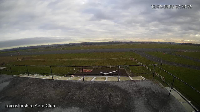 A cloudy sky is visible above a grassy airfield. In the foreground, there is a flat rooftop area with a railing. Below, an open space has a runway with a white cross marking the landing zone. To the left, a flagpole is seen with a flag. The grass around the runway is green, and there are some distant trees and landscape visible beyond the field. The time displayed on the image is 07:59:55 on December 3, 2025.