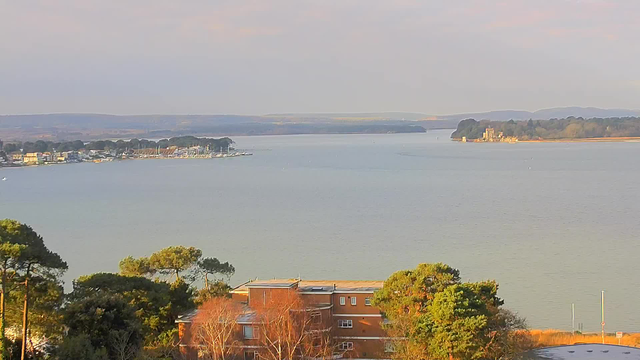 A panoramic view of a calm body of water with distant hills in the background. On the left, there is a shoreline lined with trees and buildings, including some boats docked at a marina. In the foreground, a reddish-brown building is partially visible, surrounded by greenery. The water reflects soft, muted colors of the sky, suggesting a tranquil atmosphere.