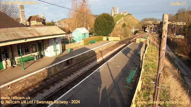 A train station scene with a platform adorned with green benches. To the left, there is a stone building with a slanted roof. In the background, a hill rises with a castle ruin at the top, surrounded by trees. The platform is mostly empty, with train tracks extending into the distance. A green sign reads "WAY OUT," and utility poles line the edge of the platform. The scene is illuminated by morning sunlight, creating long shadows.