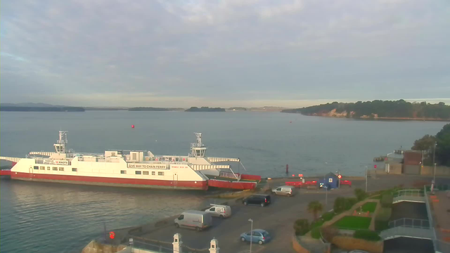 A ferry boat, painted white with red trim, is docked at a harbor on a calm body of water. The background features lush green hills and a rocky coastline under a cloudy sky. Several vehicles, including cars and a van, are parked near the ferry terminal. There are some red buoys floating in the water, and a small blue building is visible near the waterfront. The scene conveys a peaceful, maritime atmosphere.