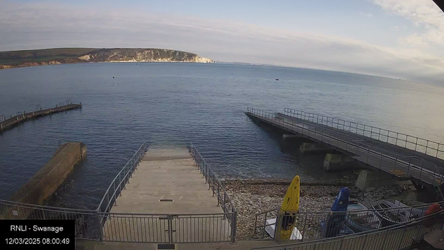 A coastal scene captured from a webcam, featuring a calm sea with a slight ripple. In the foreground, there is a concrete ramp leading down to the water, flanked by metal railings. To the right, two piers extend into the water; one is wider and has a flat surface, while the other is narrower. Two colorful boats, one yellow and one blue, are visible on the shore near the ramp. The background showcases a distant white cliff, and the sky is mostly clear with some clouds.