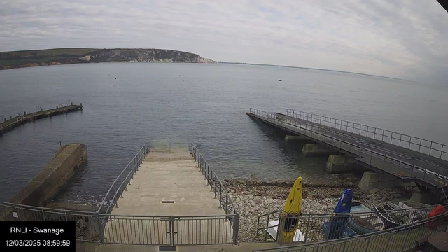 A tranquil seaside view at Swanage with calm water reflecting a cloudy sky. In the foreground, a set of concrete steps leads down to the water, flanked by railings. To the right, there is a wooden pier extending into the sea. A small storage area shows a few kayaks in yellow, blue, and red colors. The background features a rocky coastline with cliffs, and a distant landmass under an overcast sky.