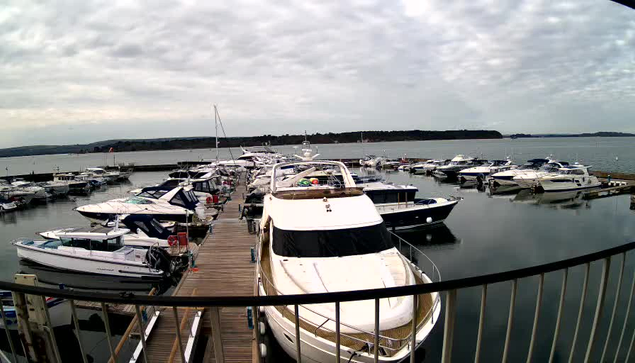 A marina scene filled with numerous boats and yachts docked at a wooden pier. The boats vary in size and style, with some featuring colorful coverings and others having sleek designs. The water is calm and reflects the cloudy sky above, creating a serene atmosphere. In the background, a tree-lined shore can be seen, with a few distant hills rising. The overall mood is peaceful and nautical.