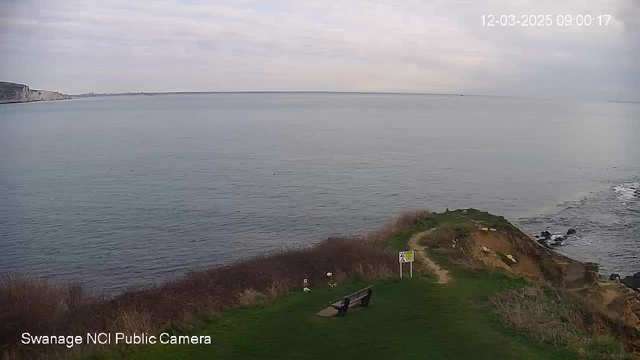 A coastal view showing a calm sea stretching to the horizon under a cloudy sky. In the foreground, there is a grassy area with a wooden bench facing the water, and a path leading to a sign nearby. The shoreline features cliffs and rocks on the right side, with some vegetation and shrubs lining the path. The scene is peaceful, with no visible people or boats.