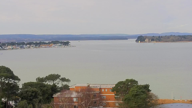 A wide view of a calm body of water reflecting soft light, with distant hills on the horizon. The shoreline features several boats and buildings, including a mix of residential and commercial structures, with trees and greenery in the foreground. There is an island in the distance, with structures that appear to be a castle or large house. The sky is overcast, giving the scene a muted atmosphere.