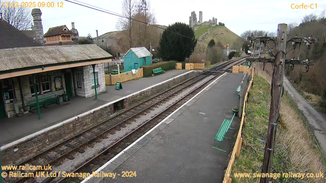 A view of a railway station with a stone building featuring a peaked roof and several windows. Green benches are placed along the platform, which is situated next to two railway tracks. In the background, there are remnants of a castle on a hillside, surrounded by trees. A blue shed and wooden fencing are visible on the station grounds. The scene is overcast, indicating it may be a chilly day.