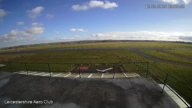 A view from a webcam overlooking the Leicestershire Aero Club. The foreground features a dark rooftop with a railing. Below, there is a grassy area with a red and white square and a white airplane silhouette marked on the ground. In the distance, a runway is visible, lined with green grass and a few trees. The sky is partly cloudy with a bright blue background. The date and time display indicate March 12, 2025, at 9:00 AM.