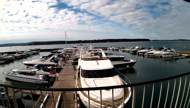 A marina filled with various boats is depicted, with a wooden dock extending into the water. Several yachts are moored along the dock, partially covered by shadows, while some are under bright sunlight. The scene is enhanced by a mixture of soft clouds scattered across the blue sky. In the background, there is a calm body of water reflecting the boats and sky, with a distant shoreline visible.