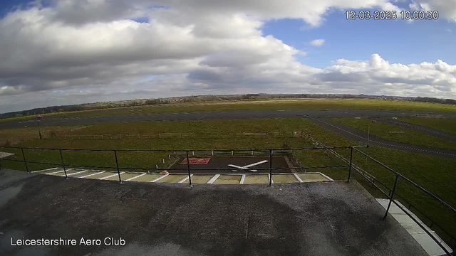 A view from a webcam overlooking a grassy airfield. The foreground features a railing and a landing pad with a white cross shape. In the background, there are clouds in a blue sky, with light and shadow across the landscape, and a mostly clear airstrip extending into the distance. The area is surrounded by a fence, with some distant trees and open fields visible. The image timestamp indicates it is March 12, 2025, at 10:00 AM.