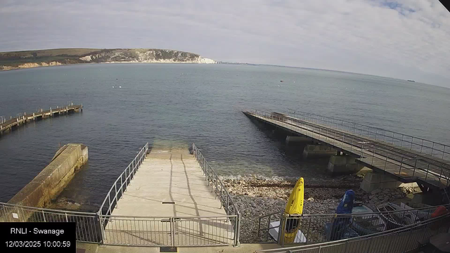 A view of a coastal area with calm blue water. In the foreground, there is a concrete ramp leading down to the shore, surrounded by a gray railing. To the right, there are two wooden piers extending into the water, with one being wider and closer to the center of the image. On the left, a curved jetty juts out into the bay. There are a few small boats visible on the water, and several mooring buoys scattered in the distance. The background features a rocky cliffside and green hills under a partly cloudy sky. A pair of yellow kayaks are secured near the ramp on the shore.