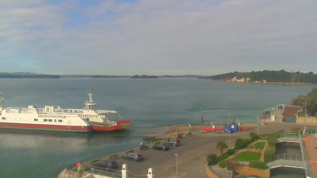 A view of a calm harbor with a large ferry docked at the pier on the left. The ferry is white with red accents and has a sign that reads "GIVE WAY TO CHAIN FERRY." Several cars are parked nearby on a lot in front of the ferry. In the background, there are small islands on the water, and a shoreline with trees and a light-colored cliff can be seen on the right. The sky is mostly blue with some clouds.