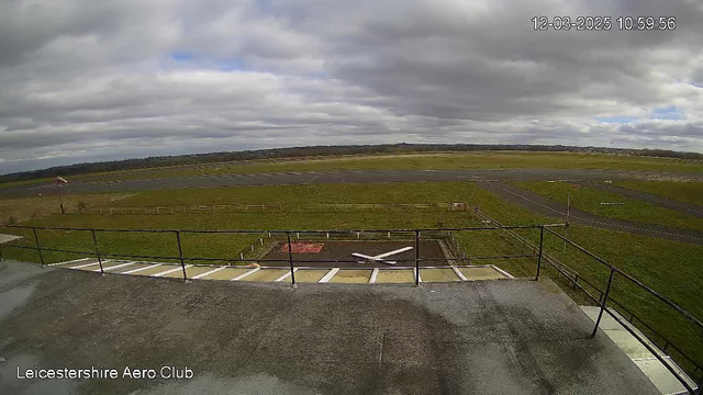 A view of an aerodrome from a webcam situated atop a building. The foreground includes a flat rooftop with a railing. Below, a green grassy area is visible, along with a tarmac runway that stretches into the distance, bordered by fences. The sky is mostly cloudy with patches of blue, and the entire scene appears calm and open. The time displayed in the corner shows 10:59:56 on March 12, 2025.