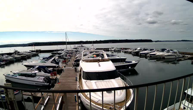 A wide view of a marina featuring numerous boats docked in calm water. The foreground includes a large white yacht with a wooden deck, while several smaller boats are visible around it. The background shows a cloudy sky and a landscape of hills or land in the distance, contributing to a tranquil atmosphere.