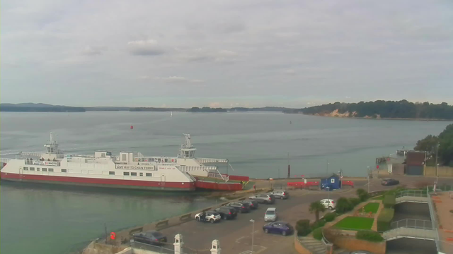 A large white ferry with a red bottom is docked at a harbor on the left side of the image. The water is calm and reflects the cloudy sky. In the background, there are green hills and a sandy shoreline. Several parked cars are visible in a parking area near the ferry, and there is a landscaped area with greenery and pathways on the right side. The scene conveys a serene coastal environment.