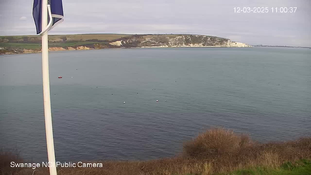 A coastal scene featuring calm, blue water with several small buoys scattered across the surface. On the left side, a white flagpole stands with a blue flag. In the background, a cliff with greenery slopes down to the water, contrasting with the white chalk cliff face. The sky is mostly cloudy, creating a muted light over the landscape. The date and time are displayed in the corner, indicating it is March 12, 2025, at 11:00 AM.