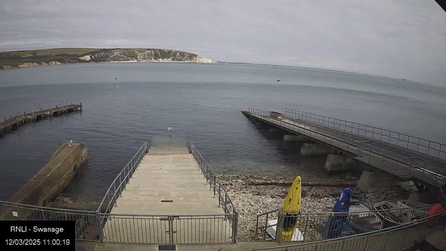 A calm coastal scene viewed from a webcam. In the foreground, there are concrete steps leading down to the water, with a metal railing along the edges. To the right, two small docks extend into the water, with one lighter-colored and one darker. On the shoreline, there are several colored kayaks, one yellow and one blue. The water is clear and reflects the gray sky, which is overcast with light clouds. In the distance, a white chalk cliff rises above the shoreline.