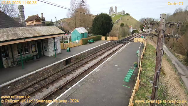 A view of a train station with platforms made of stone and asphalt. On the left, a building with a sloped roof and a chimney, with a few green benches in front. A small green shed is visible behind the building. The platform extends to the right, with train tracks running alongside. In the background, a grassy hill rises where a castle ruin is located. There are trees and a wooden fence along the platform, with a sign reading "WAY OUT" positioned on the ground. The sky is overcast.