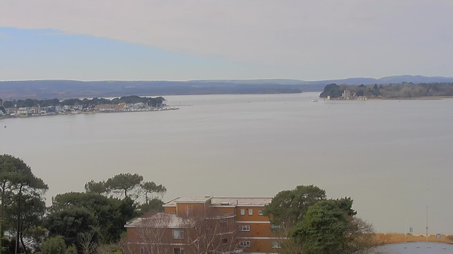 A wide view of a calm body of water with a light grey surface reflecting the sky. In the foreground, there is a brown multi-story building surrounded by trees. To the left, a marina with various boats and buildings is visible along the shore, and further in the distance, rolling hills can be seen under a light blue sky with some clouds. On the right side, a small island with a structure is situated near the water's edge. The overall scene captures a peaceful coastal landscape.