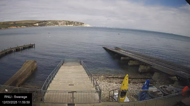 A view of a calm seaside scene featuring a wooden jetty extending into the water on the left, a concrete ramp leading down to the sea in the center, and a rocky shore. Two brightly colored kayaks, one yellow and one blue, are positioned on the right side near the water's edge. The sky is partly cloudy with gentle waves visible on the surface of the water, while distant cliffs can be seen in the background.